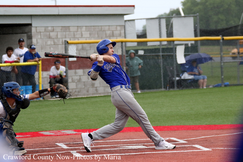 Youth baseball batter in a blue uniform swings at the ball from a right-handed stance as the catcher in black gear crouches behind home plate.