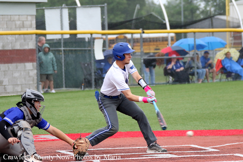 Baseball batter in white and blue swings at a pitch as a catcher in blue gear prepares behind him on a rain-soaked field with spectators under umbrellas in the background.