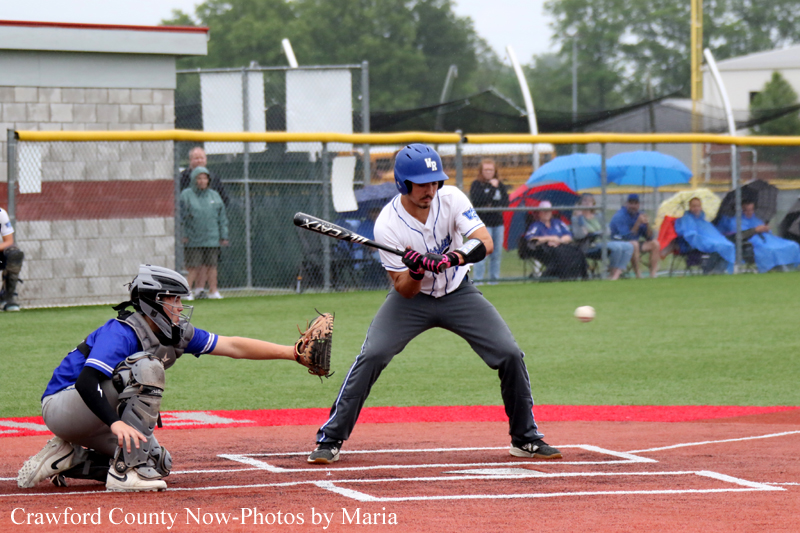 Baseball batter in a blue helmet swings at a pitch as the catcher crouches behind home plate on a red-dirt field.