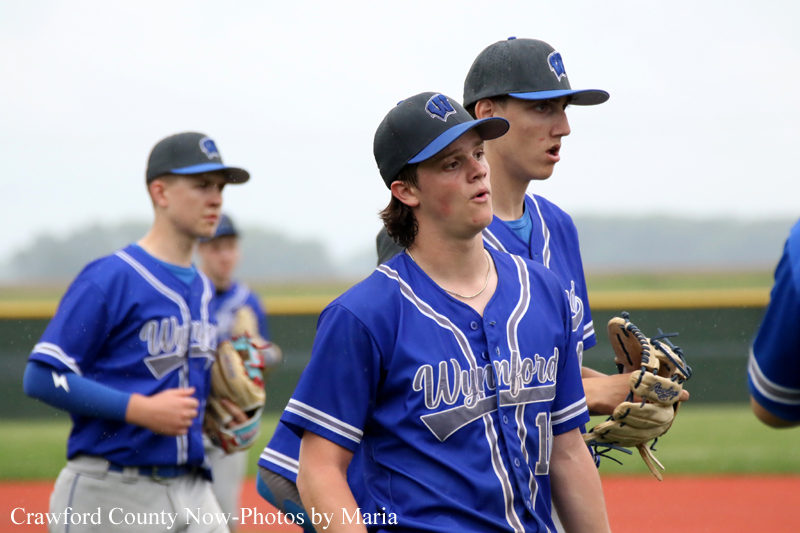 Three Wynford baseball players in blue uniforms walk onto the field, foreground player wearing a cap and glove and looking ahead.