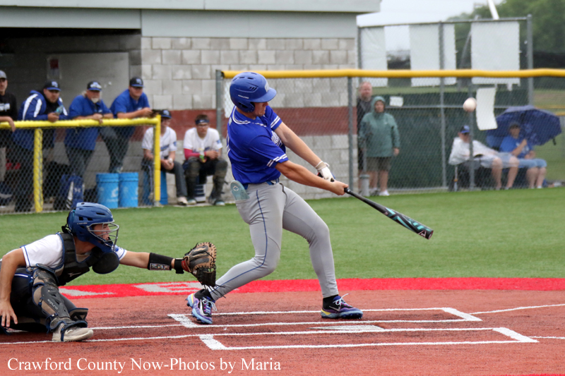 Baseball batter in blue uniform swings the bat as the catcher in black gear lunges toward home plate.