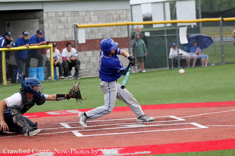 Youth baseball game: a batter in a blue uniform swings at a pitch as the catcher in black gear crouches behind home plate; spectators watch from the fence.