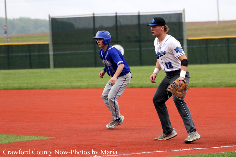 Two young baseball players sprint along the infield dirt toward base, one in a blue uniform with helmet and the other in a white uniform with a glove, focused on the play.