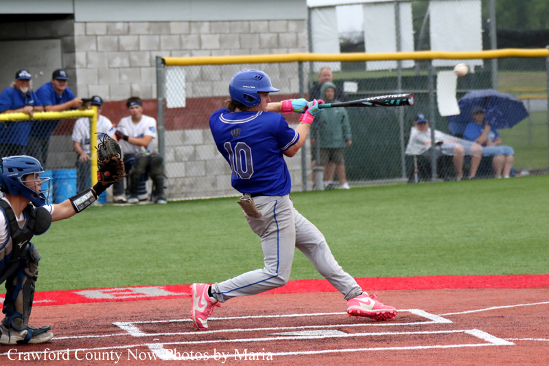 Baseball batter in a blue uniform swings at a pitch with pink cleats, as the catcher crouches behind home plate and spectators watch beyond the fence.