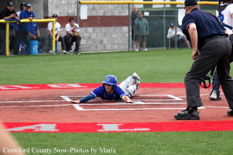 Blue-uniformed baseball player slides headfirst into home plate as umpire watches on a grassy field with spectators in the background.