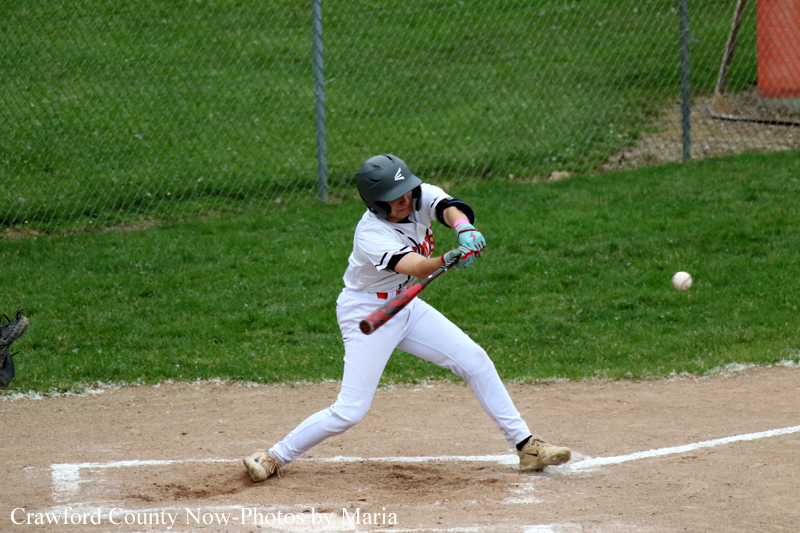 Baseball batter in white uniform swings at a pitched ball near home plate on a dirt field, grassy outfield in the background.