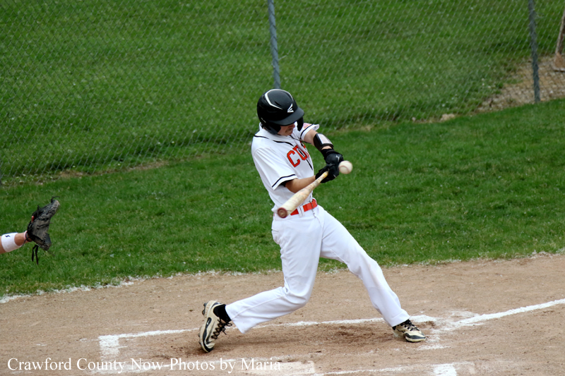 Baseball batter in a white uniform swings at a pitched ball on the dirt batter's box with a black helmet and bat mid-swing.