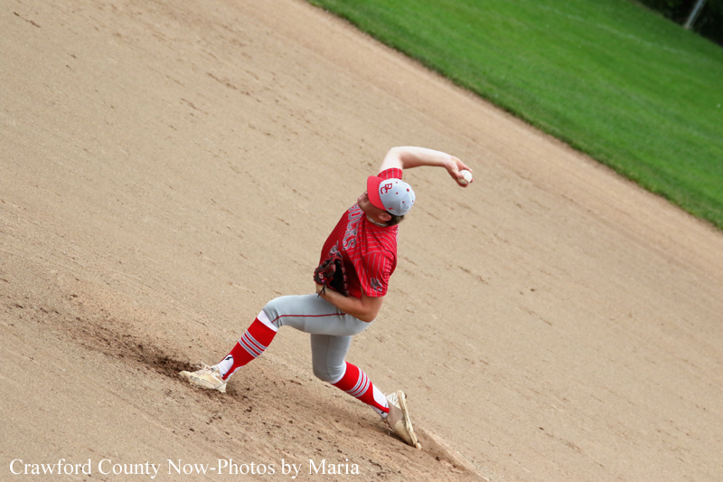 Youth baseball pitcher in a red uniform throws a pitch from the dirt mound with grass in the background to the right.