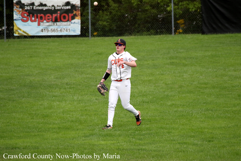 Baseball player in a white uniform watches a ball in the air with a glove ready on a grassy field with a banner in the background.