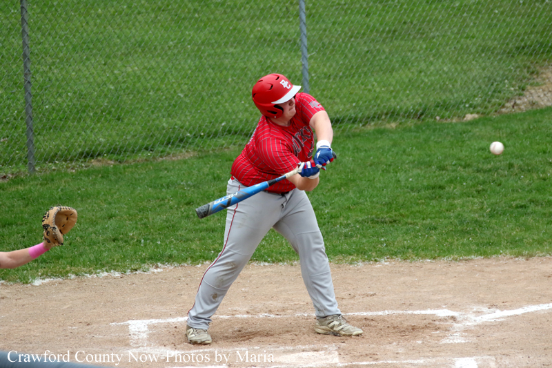 Baseball batter in red uniform swings at a pitch during a game on a dirt field with a catcher’s glove visible to the left