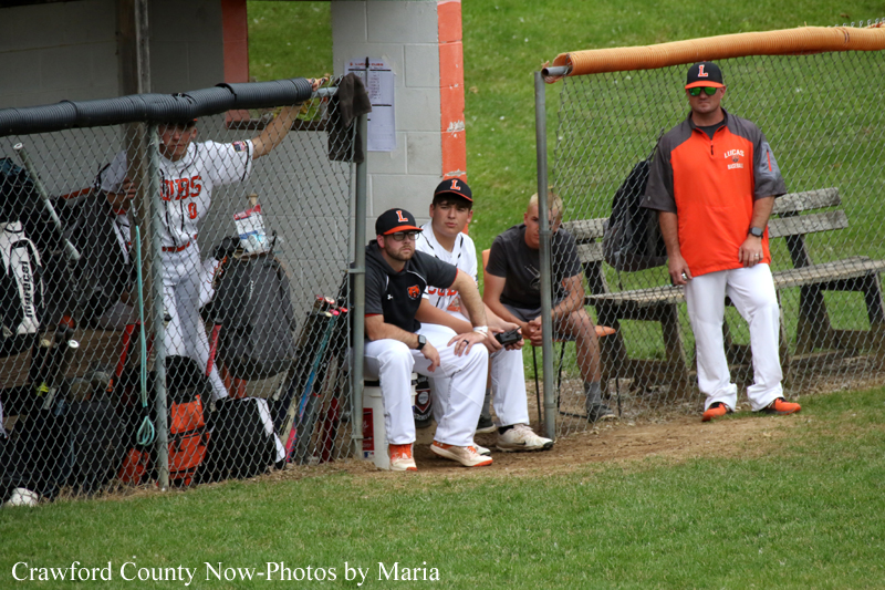 Baseball players sit in a dugout behind a chain-link fence, with a coach standing outside in an orange jacket and white pants.