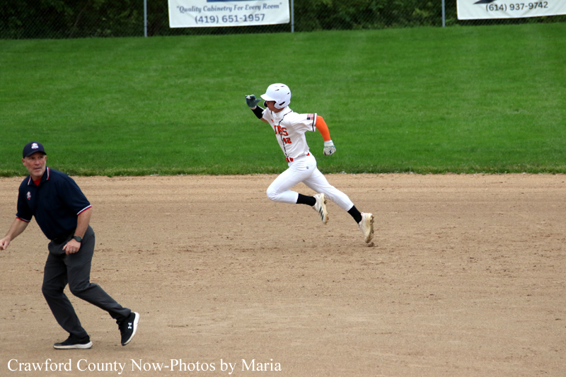 Baseball runner in white uniform sprinting across the infield dirt, with an umpire nearby on the left and green outfield in the background.