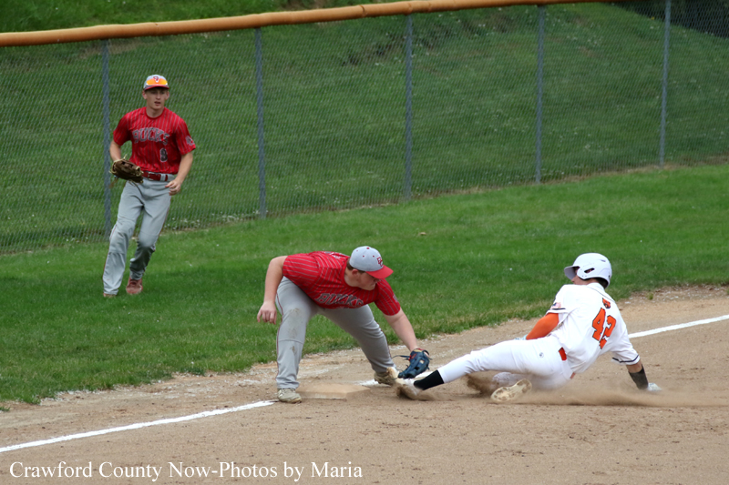 Baseball play at the infield: a red-jersey fielder reaches with his glove as a white-uniform runner slides headfirst into the base, while another player watches from the grass behind the fence.
