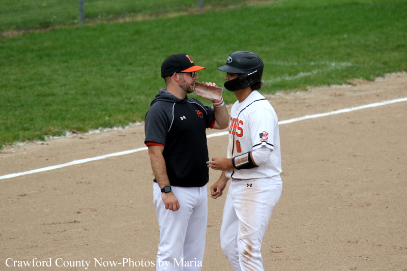 Two baseball players stand on the infield dirt near the baselines, one in a black hoodie and orange cap offering a drink to a teammate in a white uniform and helmet.