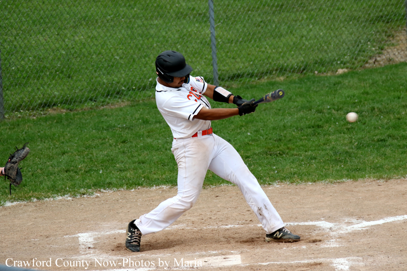Baseball batter in a white uniform swings at a pitch on a dirt infield with green grass in the background.