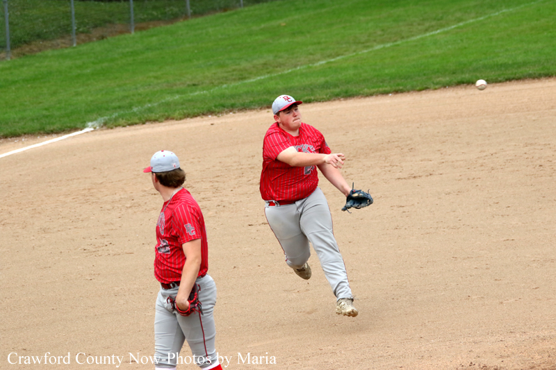 Two baseball players in red uniforms on a dirt infield; one pitcher is mid-throw while a teammate watches nearby.