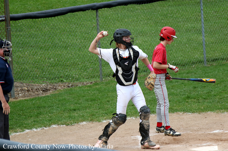 A baseball catcher in white uniform and protective gear throws a ball while a batter in red waits nearby on the field with a bat on the ground behind them.