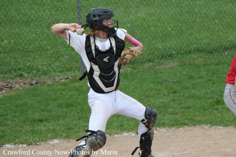 Youth baseball catcher in white uniform and protective gear throws a ball on a grassy field with a chain-link fence in the background.
