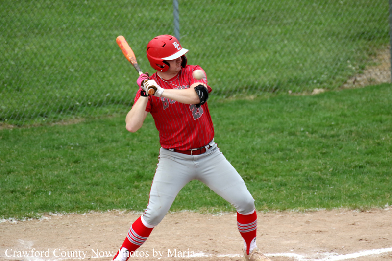 Baseball player in red uniform swinging a bat at a pitch on a dusty field with a green outfield in the background.