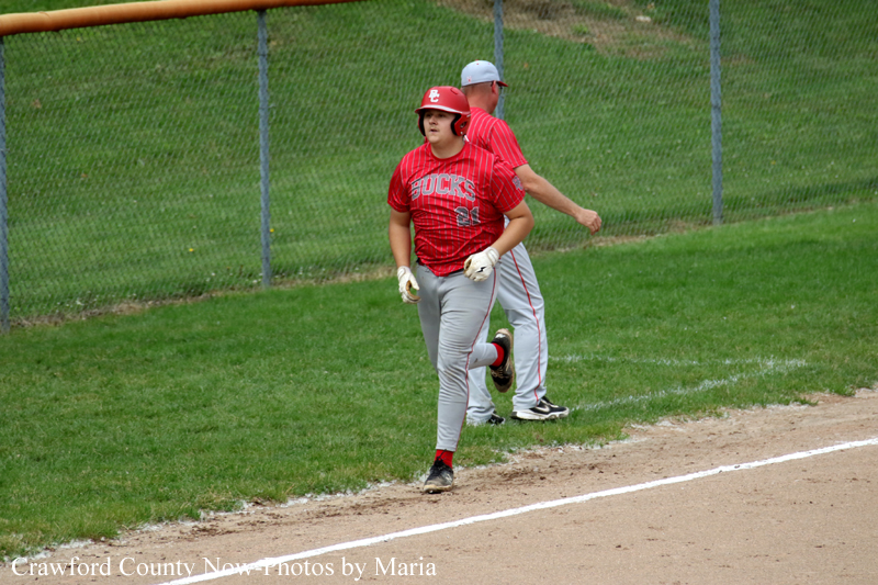 Baseball player in a red uniform runs along the first-base line while a coach watches in the background on a grassy field.