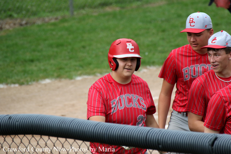 Three baseball players in red BUCKS uniforms stand near a fence on a dirt field, one wearing a red helmet with BC.