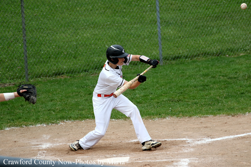 Baseball batter in a white uniform swings at a pitch on a dirt field with a catcher’s glove visible to the left and a chain-link fence in the background.