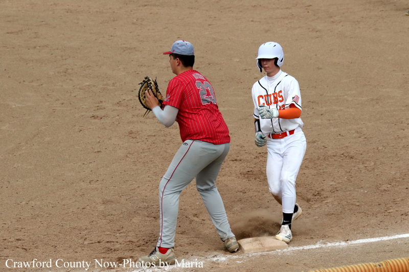 Baseball play at first base: fielder in red jersey reaching with his glove as a Cubs baserunner in white nears the bag on a dirt field