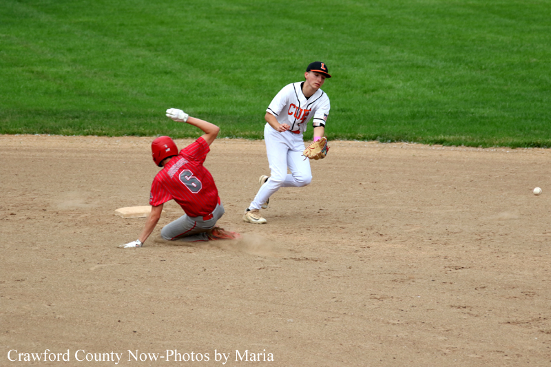 Baseball action: runner slides into a base as a fielder in white chases the play, ball nearby in the dirt.