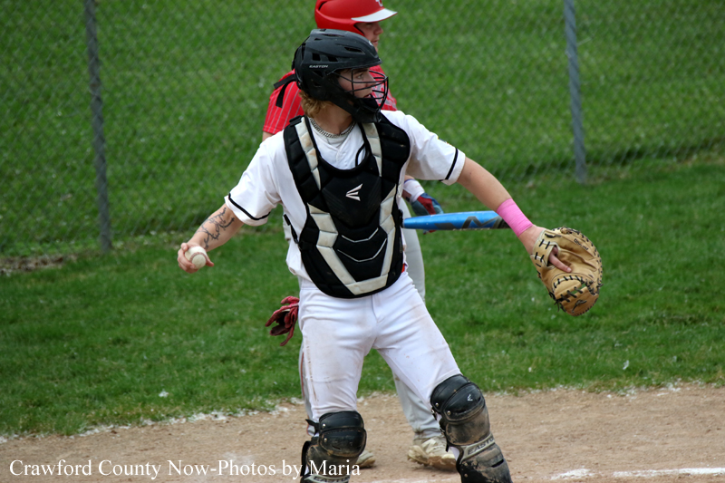 Youth baseball catcher in a white uniform with black protective gear, glove ready on a dirt field during a pitch.