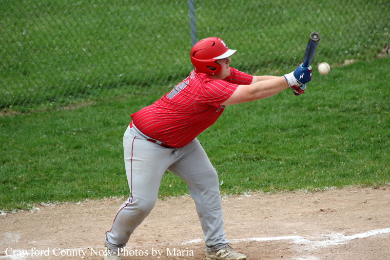 Baseball player in a red jersey and helmet swings a bat at a pitched ball on a dirt infield with a chain-link fence in the background.