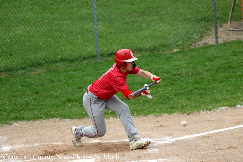 Young baseball player in a red uniform and helmet swings a bat at a pitch near home plate on a dirt infield.