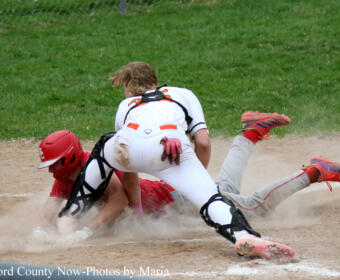 Baseball action: a catcher in white gear slides while a runner in red starts to collide at a dusty base, kicking up dirt on the infield.