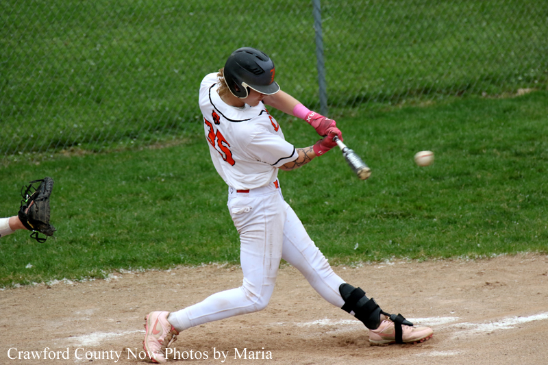 Female baseball player in a white uniform swings at a pitched ball during a game at the plate, pink gloves on.