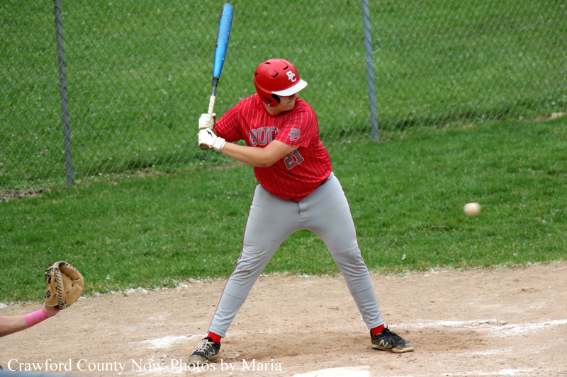Young baseball batter in a red uniform and helmet swings a blue bat at home plate, with a fielder’s glove visible in the foreground.