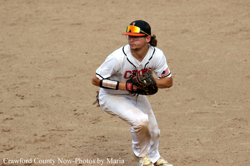 Baseball infielder in a white uniform crouches with glove, ready to field on a dirt infield.