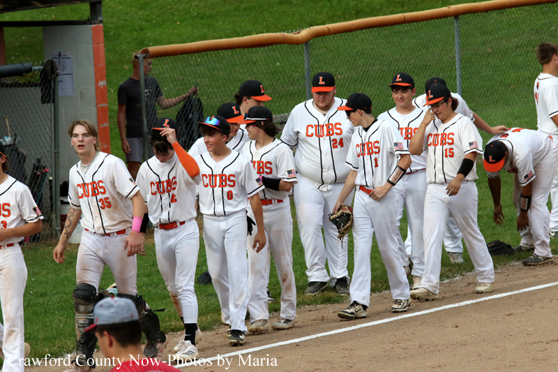 Youth baseball team in white uniforms with 'CUBS' on chest walk along the dugout fence.