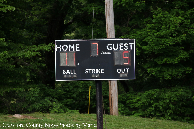 Outdoor baseball scoreboard on a metal post showing Home 11, Guest 5, with Ball, Strike, and Out indicators in a grassy field with trees in the background.