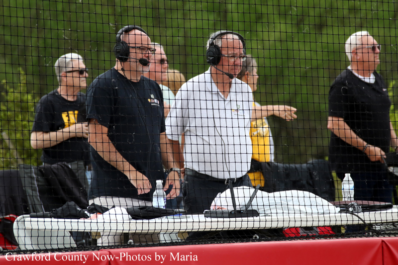 Broadcast team with headsets and microphones at a game announcer table behind a protective net.