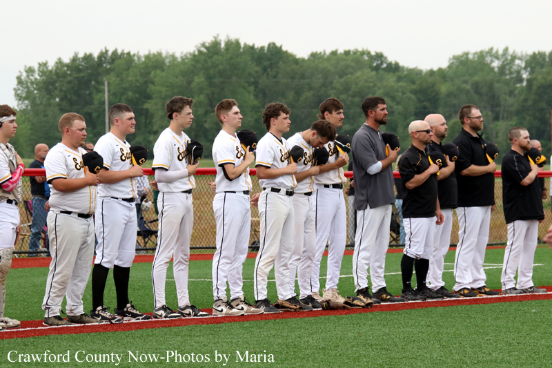 Baseball players in white uniforms stand in a line along the field with hands over their hearts during the national anthem.