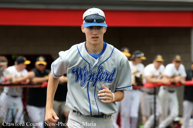 Baseball player in a gray blue-trimmed uniform running toward the camera, wearing a white cap with blue brim and sunglasses on top, in a dugout setting with teammates in the background.