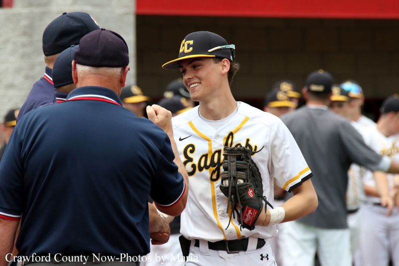 Baseball player in a white Eagles uniform shakes hands with a coach, smiling as teammates watch in the background.