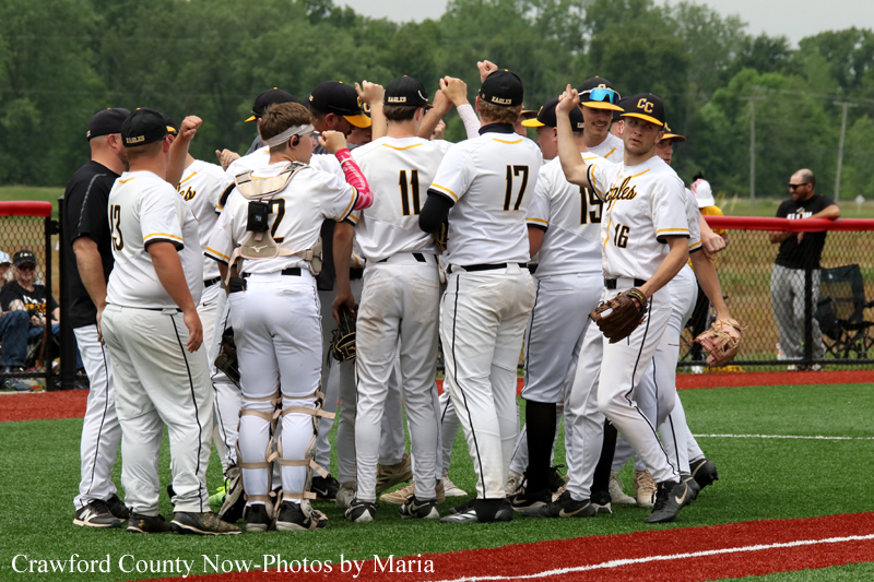 Baseball players in white uniforms huddle on a green field, arms raised in celebration near a chain-link fence.