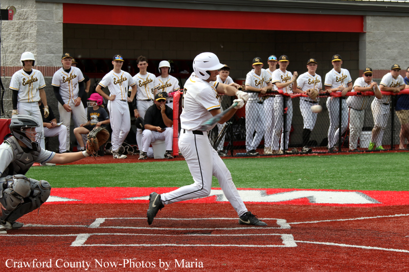 Baseball batter in a white uniform swings at a pitch, with the catcher and umpire behind home plate and teammates watching from the dugout.