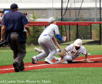 Baseball play at a bag: a runner slides while a fielder reaches for the ball; an umpire stands nearby as teammates watch in the background.