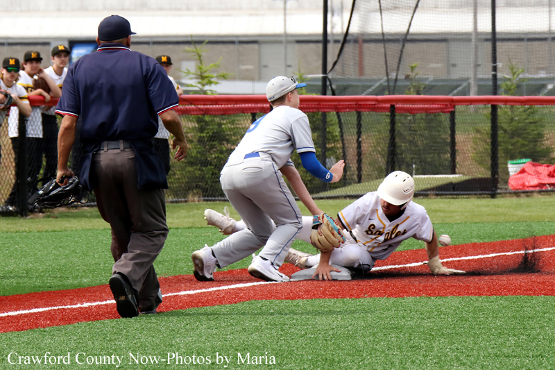 Baseball play at a bag: a runner slides while a fielder reaches for the ball; an umpire stands nearby as teammates watch in the background.
