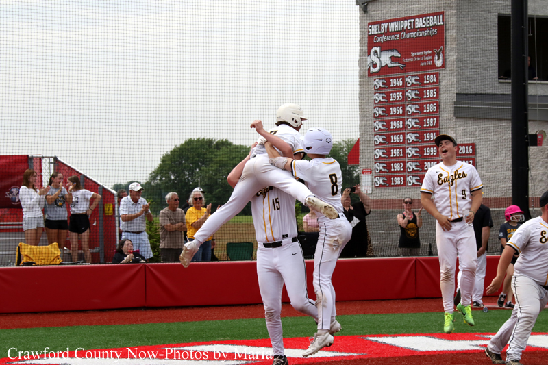 Two players in white-and-gold uniforms leap into the air in celebration at home plate, with teammates and spectators behind a red barrier watching.