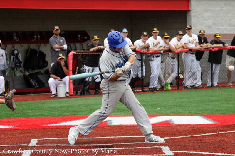 Baseball batter in gray uniform and blue helmet swings at a pitch at home plate, with teammates watching from the dugout behind a chain-link fence.