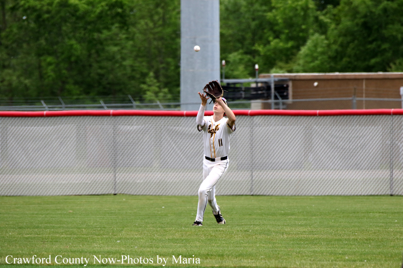 Baseball player in a white uniform reaches up to catch a fly ball with a glove in the outfield, fence in back.