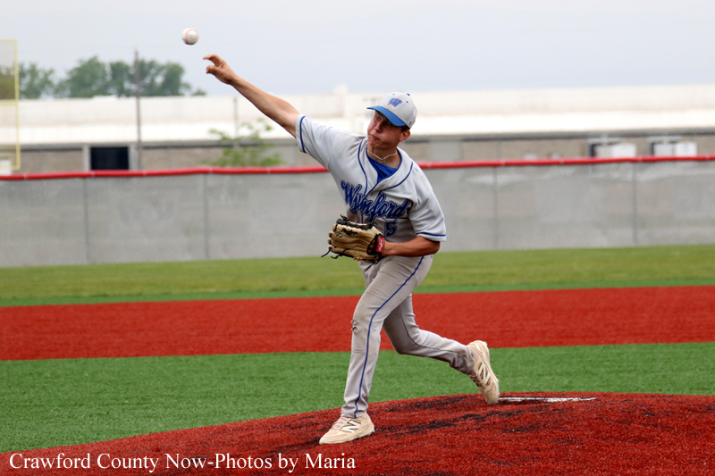 Baseball pitcher in a gray uniform throwing a pitch on the mound during a game, with a chain-link fence in the background.