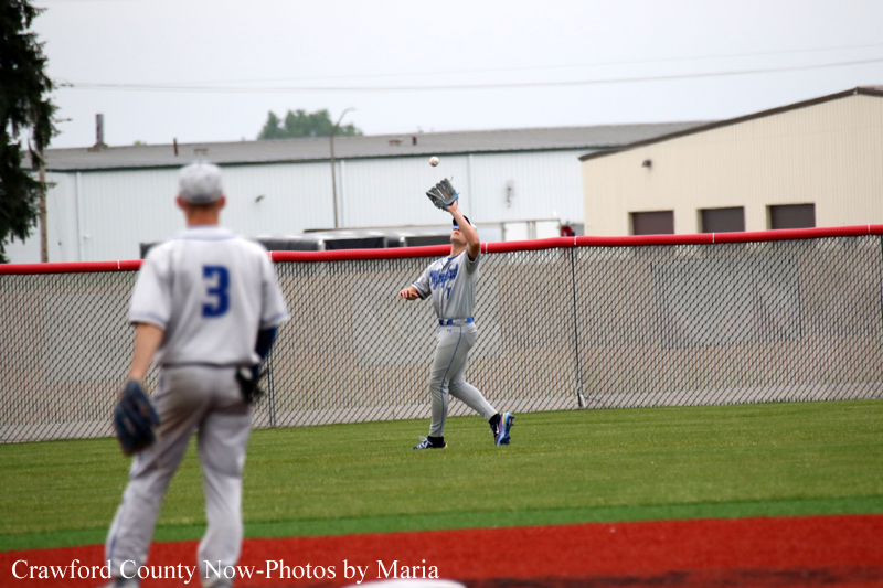 Two baseball players on a grassy field near a chain-link fence; a gray-uniform fielder reaches up with his glove as a ball is tossed overhead, while another player in the foreground watches (back to camera).